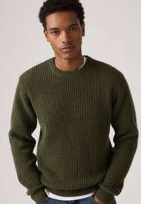 Donkergroene gebreide trui met een gestructureerd wafelpatroon, ronde halslijn en geribbelde manchetten. G gedragen over een wit shirt, casual pasvorm.