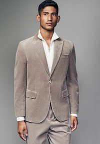 Helles taupe Cord-Blazer mit einem einzelnen Knopf, fallenden Revers und Vordertaschen, kombiniert mit einem weißen, durchgehend mit Reißverschluss versehenen Kleidungsstück darunter.