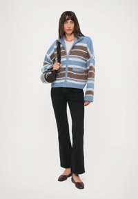 Mujer con un suéter de cremallera a rayas azules y marrones, jeans flacos negros, zapatos planos burdeos y llevando un bolso cilíndrico negro sobre el hombro.