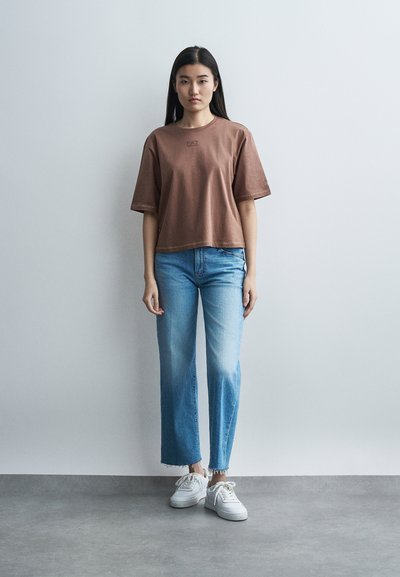 Μία γυναίκα στέκεται μπροστά σε έναν απλό τοίχο φορώντας καφέ φαρδιά μπλούζα, ανοιχτόχρωμο μπλε τζιν παντελόνι κατεβασμένο με frayed τελειώματα και λευκά αθλητικά παπούτσια.