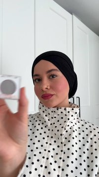Heldere glazen container met grijs cosmetisch product. Model draagt een zwarte hijab en een witte jas met stippen, en houdt de container naar de camera gericht.
