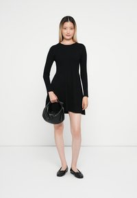 Vestido preto canelado com mangas longas e saia rodada. O modelo segura uma pequena mala preta. Ela usa sapatos planos pretos com laços. Textura suave.