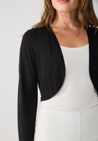 Black cropped cardigan with long sleeves, made of lightweight material. Worn over a white top, featuring a smooth texture and open front design.