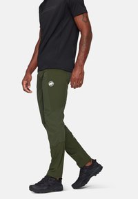 Olijfgroene outdoor broek met een slim fit, voorzien van een logopatch, zijzakken en contrasterende stofpanelen, gecombineerd met zwarte schoenen en een shirt.
