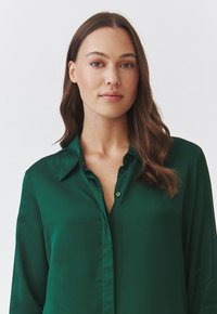 Blusa di raso verde con colletto classico, chiusura anteriore con bottoni, maniche lunghe e una superficie liscia e lucida.