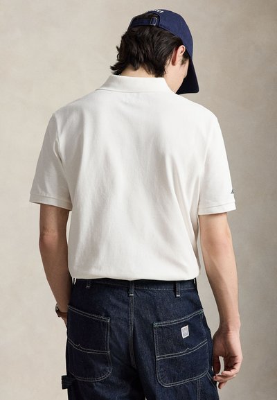 Άτομο που φοράει άσπρο πόλο μπλουζάκι με κοντό μανίκι και σκούρο μπλε τζιν παντελόνι, με την πλάτη γυρισμένη, φορώντας μπλε σκούφο μπέιζμπολ προς τα πίσω.