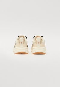 Sneaker beige con una toma superiore liscia e resistente, dettagli neri e una suola bianca spessa. Presenta un'etichetta sul tallone per indossarle facilmente.