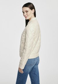 Pikowana kremowa kurtka z wysokim kołnierzem, prążkowanymi mankietami i dołem, zestawiona z niebieskimi dżinsami. Posiada lekkie, błyszczące wykończenie.