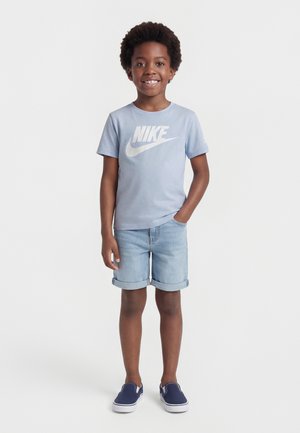Ung gutt står og smiler, iført en lyseblå Nike t-skjorte, oppbrettede denimshorts og marineblå slip-on sko.