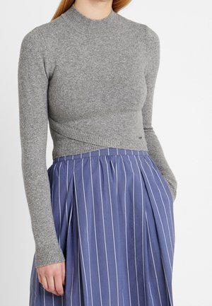 Vrouw draagt een aansluitende grijze trui met lange mouwen en een hoge tailleblauwe rok met witte verticale strepen, hand in de zak.