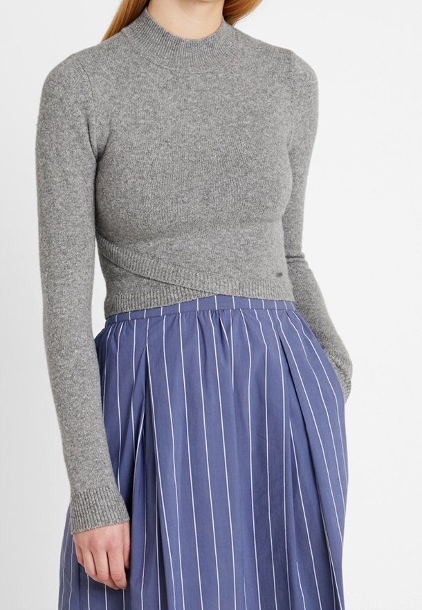 Femme portant un pull gris ajusté à manches longues et une jupe bleue taille haute avec des rayures blanches verticales, main dans la poche.