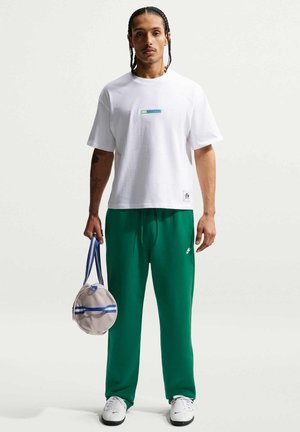 Man met gevlochten haar, draagt een wit T-shirt en groene joggingbroek, houdt een ronde grijze tas met blauwe bandjes vast, staand tegen een witte achtergrond.