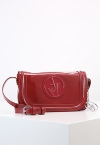 Borsa a spalla rossa lucida con tracolla regolabile, logo AJ in rilievo sul davanti e ciondolo in argento con logo AJ appeso di lato.