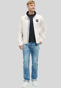 Uomo che guarda a destra, indossando una giacca bianca con zip, una maglietta nera, jeans blu e sneaker bianche, in piedi su uno sfondo grigio chiaro uniforme.
