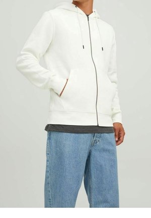 Person trägt einen weißen Kapuzenpullover mit Reißverschluss, Fronttaschen und Kordelzug, kombiniert mit einem dunklen T-Shirt und hellblauen Jeans.