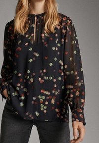 Kvinna i en svart blus med ett multikolorert abstrakt prickmönster, långa genomskinliga ärmar och en nyckelhålsringning med knytning.