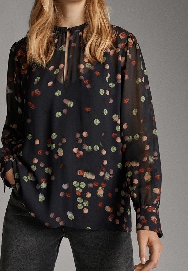 Kvinna i en svart blus med ett multikolorert abstrakt prickmönster, långa genomskinliga ärmar och en nyckelhålsringning med knytning.