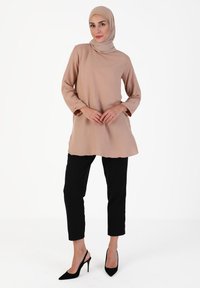 Langærmet, beige tunika med høj hals, parret med sorte, slanke bukser og sorte hæle. Glat stof, afslappet silhuet.
