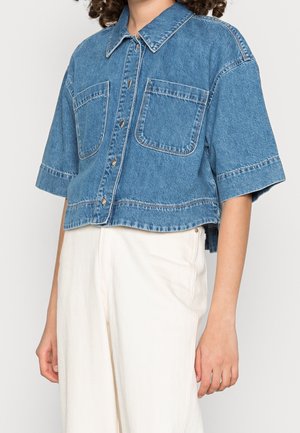 Persoon draagt een cropped blauwe denim blouse met knopen vooraan en brede korte mouwen, gecombineerd met hooggetailleerde crème broek.