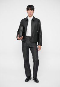 Chaqueta de cuero negra con cierre de cremallera, camisa blanca con cuello, pantalones de mezclilla oscuros y zapatos negros. Sosteniendo un bolso rectangular negro.