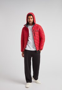 Pelliccia rossa imbottita con cappuccio, chiusura con zip e fodera grigia. Indossata sopra una camicia bianca e abbinata a pantaloni neri e sneakers bianche.