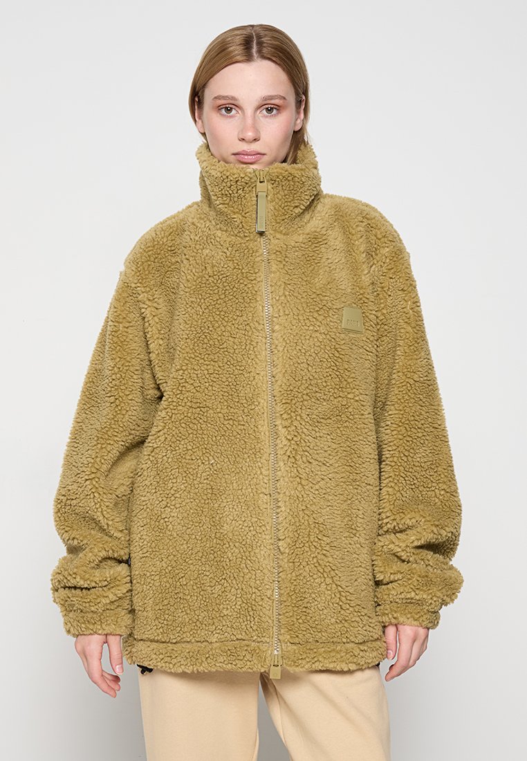 Veste zippée en polaire de couleur brun clair, avec un col montant, une coupe oversize et un tissu texturé, arborant un logo de la marque sur la poitrine.