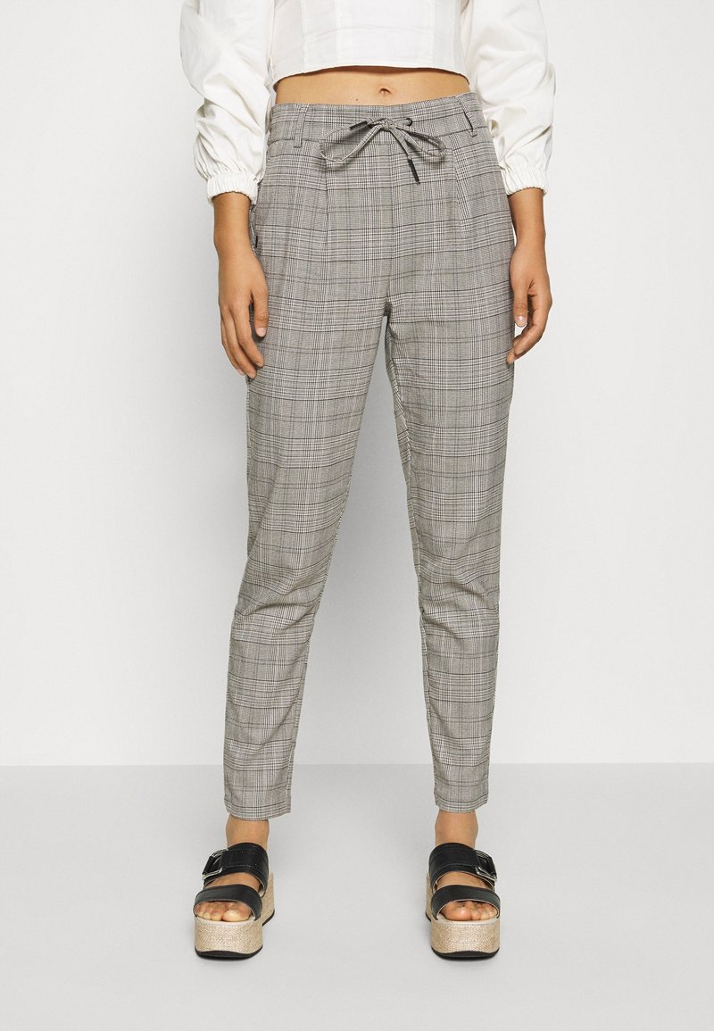 Une femme porte un pantalon à taille haute en tissu écossais gris avec une taille à cordon, ainsi que des sandales plateforme noires, associé à un chemisier blanc court.