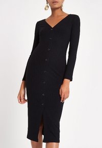 Robe noire côtelée à manches longues, col en V, fermeture à boutons sur le devant et fente latérale. Texture douce, coupe cintrée.