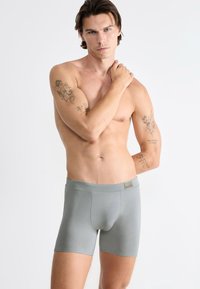Graue Boxer-Briefs aus glattem Material, mit einem breiten Bund und einer engen Passform. Das Modell posiert mit einer Hand auf seiner Schulter.