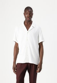 White short-sleeve button-up shirt with a relaxed fit, featuring a notched collar, textured fabric, and contrasting buttons. Worn with dark pants.