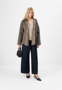 Vrouw in een bruin fleece jack, beige gebreide trui, wijdere donkere spijkerbroek, zwarte slingback hakken, met een kleine zwarte handtas.