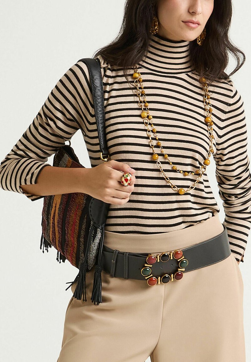 Gestreifter Rollkragenpullover in Beige und Schwarz, beidseitige beige Hose, schwarzer Gürtel mit farbenfrohen Steinelementen, Perlenkette und bunte Umhängetasche.