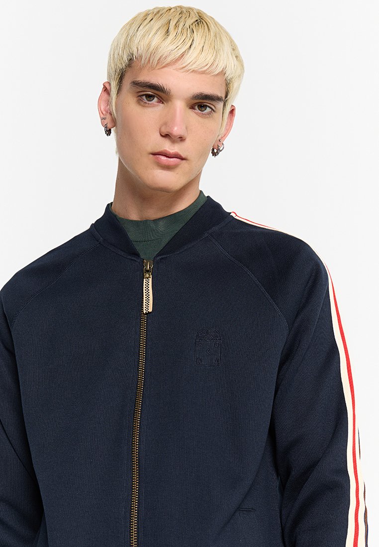 Junge Person mit kurzen blonden Haaren, die eine navyfarbene Jacke mit Reißverschluss und roten und weißen Streifen an den Ärmeln sowie silbernen Creolen trägt.