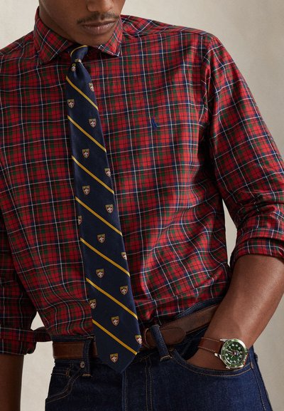 Czerwona i zielona koszula w kratę z granatowym krawatem w żółte paski i herby. Brązowy skórzany pasek i zielony zegarek na nadgarstku.