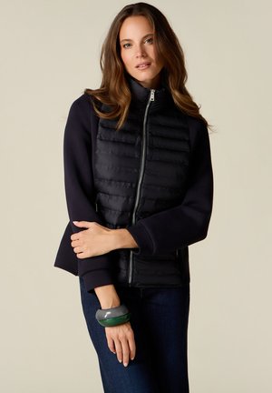 Kvinde med brunt hår iført en sort foret jakke med lynlås og mørke jeans, holder den ene arm over taljen, lysebeige baggrund.