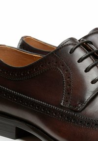 Braune Leder-Brogue-Schuhe mit filigranen Perforationen, glatter Textur, abgerundetem Zehenbereich und schwarzen Schnürsenkeln vor hellbraunem Innenfutter.