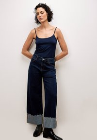 Navy Tanktop mit dünnen Trägern, kombiniert mit weiten dunklen Jeans aus Denim mit fransigen Abschlüssen und kontrastierenden hellen Denim-Akzenten. Schwarze Stiefel.