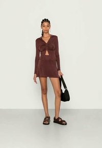 Conjunto dois peças em bordeaux, apresentando uma blusa ajustada de manga longa com um recorte frontal e uma saia mini correspondente; complementado com uma bolsa preta e sandálias.