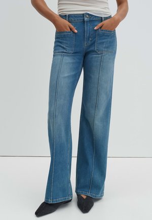 Person trägt hochtaillierte, weit geschnittene blaue Jeans mit aufgesetzten Vordertaschen, weißes Oberteil und schwarze spitze Schuhe, steht mit den Händen in den Taschen.