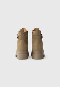 Καφέ σουέντ μποτάκια μέχρι τον αστράγαλο με ελαφρώς στρογγυλή μύτη και χοντρό τακούνι. Περιλαμβάνει λουρί με λεπτομέρεια κούμπωμα στον αστράγαλο.