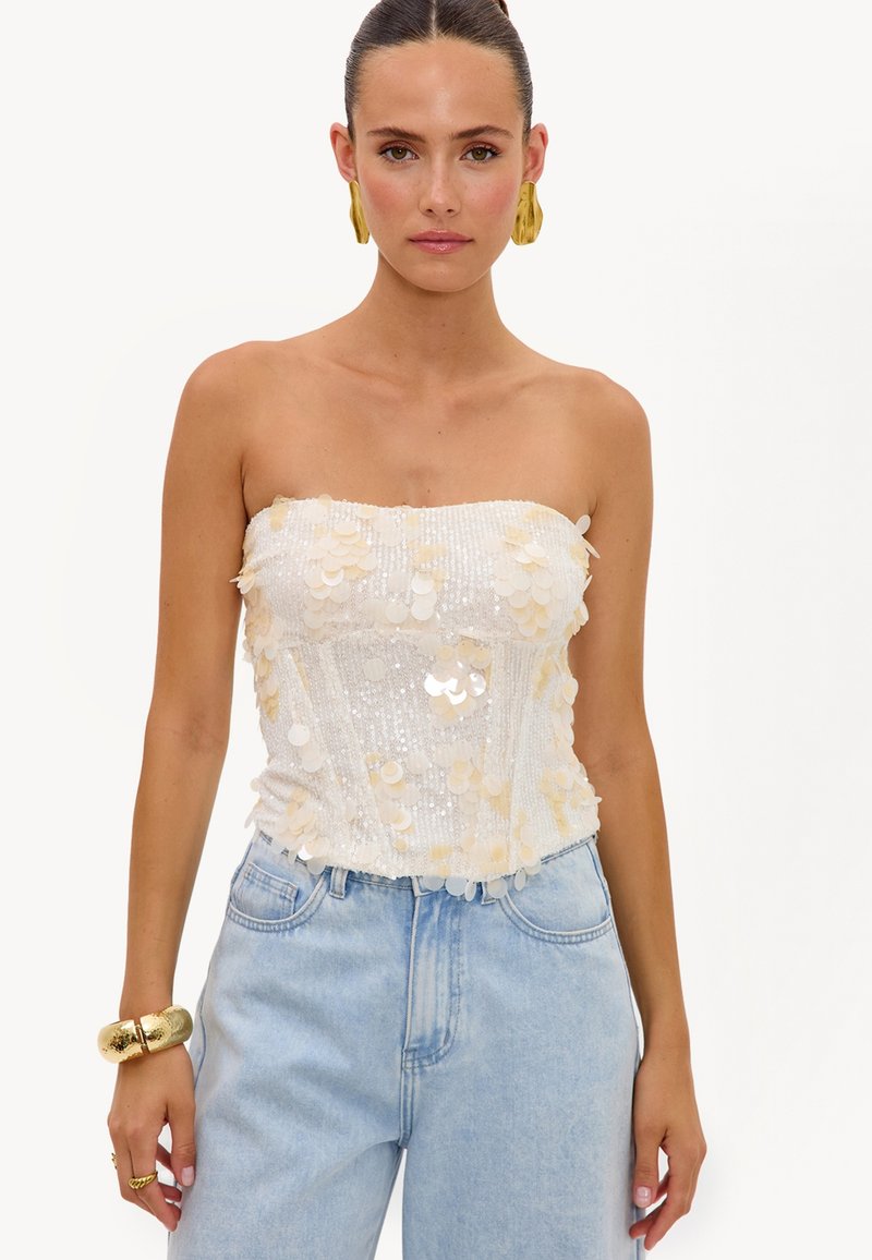 Femme portant un haut bustier blanc à sequins et un jean bleu clair avec des boucles d'oreilles, un bracelet et des bagues dorés sur un fond blanc uni.