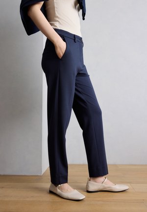 Persoană purtând pantaloni bleumarin, top bej și pantofi Mary Jane bej, stând cu o mână în buzunar pe podea din lemn.