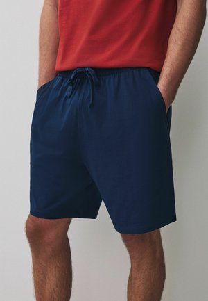Mann trägt dunkelblaue Shorts mit Kordelzug und ein rotes T-Shirt, die Hände in den Taschen, vor einem einfarbigen Hintergrund.