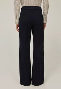 Persona che indossa pantaloni neri a vita alta e a gamba larga, insieme a una camicia a righe beige e bianca a maniche lunghe, vista da dietro.