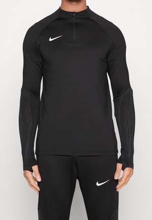 Mężczyzna w czarnym sportowym topie Nike z długim rękawem i zamkiem do ćwiartki oraz dopasowanych czarnych spodniach Nike z logo na lewym udzie.