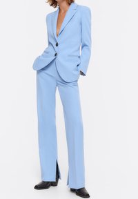 Woman wearing a light blue tailored suit with notch lapel blazer and matching wide-leg trousers with front slits, paired with black square-toe boots.