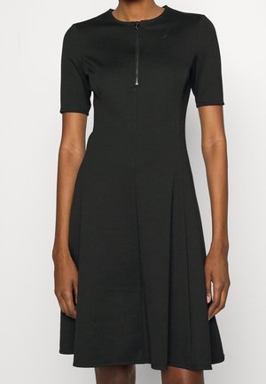 Mujer con un vestido negro hasta la rodilla, de manga corta y cremallera frontal desde el escote hasta la mitad del pecho.