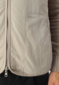 Helle graue, leichte Weste mit strukturierter Oberfläche, Vorderreißverschluss, Seitentasche und dezentem "Barbour"-Logo. Weiche Kanten und minimalistisches Design.