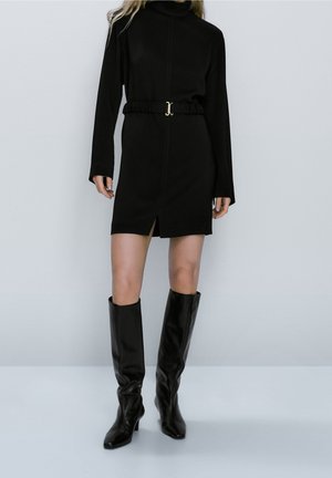 Kvinde iført en sort langærmet minikjole med taljebælte og sorte knæhøje støvler med hæl, stående foran en ensfarvet baggrund.