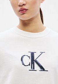 Valge puuvillane T-särk, millel on eristuv "CK" logo sinistest helmestest tikandina. Kaelus on ümara lõikega ja lühikeste varrukatega.